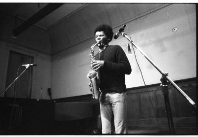The musician stands straight up lifting the saxophone with their torso. They wince as they push air through the instrument's reed. 