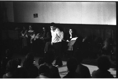 A black and white photo showing Joel Chadabe surrounded by seated audience members in the Grand Luxe Hall as he stands with his left arm outstretched, playing a theremin.