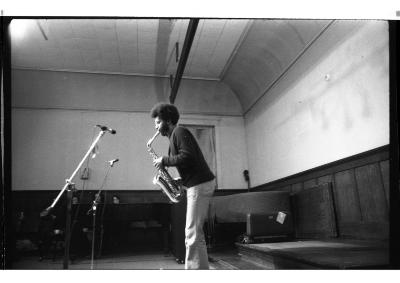 A side angle of the artist performing. Their head leans forward as their hips lean back. The saxophone follows suit. 