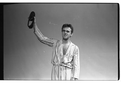 A waist-up image of a man holding a slipper with his right hand. He is wearing a matching striped shirt and pants, and his shirt is unbuttoned. His gaze looks off to the left of the image. A projector screen is in the background. The image is in black and white.
