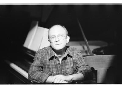 A man wearing aviator glasses and a plaid button up sits beneath a bright spotlight, smiling softly. He has a receding hairline and holds his hands together near his chest. Behind him is a beautiful grand piano. 
