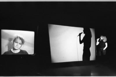 A woman stands in front of a projector screen while speaking into a wired microphone. She turns to face a monitor that displays a portrait of her tilting her head.