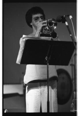 A trombonist with an afro and aviator glasses stands behind a scoreholder, looking down as he plays. He wears a light-colored long-sleeve top and white trousers. 
