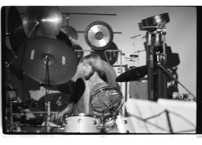 A blurry shot of a drummer surrounded by an elaborate drum set with various cymbals towering high above his head. The drummer has a bowl cut and facial hair, his head turned to the side as if in motion. 
