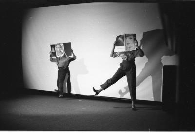 Two women stand on a spotlight-lit stage, wearing trousers, dress shirts and heels. They each hold a magazine up in front of their faces, replacing their heads with those of glammed-up models. Each woman raises one leg up as if in a choreographed dance.
