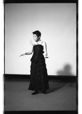 Marsha Paludan stands in front of a large blank screen. A feather is tucked into the bodice of their long, formal gown with thin straps and layered detailing. They address their attention to the ground and hold both hands out in a graceful gesture.