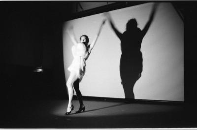 A woman wearing dark heels and a short white dress stands beneath a spotlight with one leg forward and both arms waving high up in the air. Two balloons are stuffed beneath her dress, imitating breasts. A large, imposing shadow is cast on the white screen behind her. 
