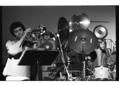 A trombonist with a small, dark afro tilts his head as he looks at his score holder, his tongue sticking out of his mouth. To his right is an elaborate white drum set with various cymbals, standing taller than the trombonist himself. A drummer with a bowl cut and facial hair sits behind the set.
