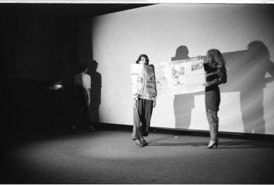 Three women stand on a stage, lit by a spotlight. One woman appears to be wrapping – or perhaps unwrapping – another woman in a scroll of newspaper. At the far edge of the spotlight, the third woman stands with her back to the camera. 
