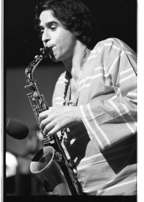 Dutch-angle shot of a dark-haired saxophonist wearing a light-colored striped long-sleeve. The artist has thick eyebrows and hooded eyes, and appears quite nonchalant. 
