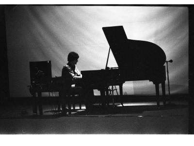 A back lit long-shot image depicting a man playing the grand piano. The backdrop behind the man ripples down and there is a computer interface instrument next to him.