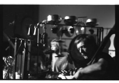 A man with short-cropped hair and a mustache sits down as he plays the bass. Between the bassist and another, longer-haired musician is an elaborate set up of cymbals and bells.
