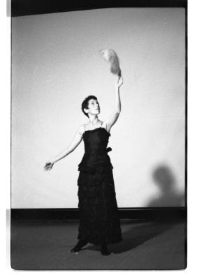 Marsha Paludan stands with their left hand raised, holding a large feather in the air, in front of a blank screen where their shadow is reflected. Their hair is closely cropped to their face. They wear a long, formal gown with thin straps and layered detailing.