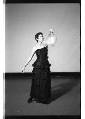 Marsha Paludan stands in front of a large blank screen. They address their attention to a feather they hold in their left hand. Their hair is closely cropped to their face. They wear a long, formal gown with thin straps and layered detailing.