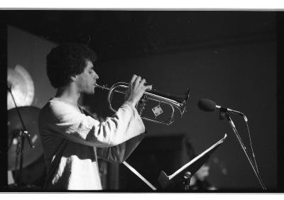 Side-profile of a trombonist standing behind a score holder and a microphone. He has a dark afro, wears a light-colored linen long-sleeve, and shuts his eyes as he plays. We can see the vague shape of drum cymbals behind him.
