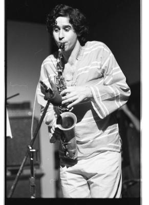 A dark-haired saxophonist wears a bell-sleeved striped top and light colored trousers as he plays his instrument. A microphone is pointed at the bell of his saxophone. 
