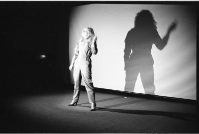 A blonde woman dressed in a long-sleeve business jumpsuit and heels stands in the center of a stage with one arm raised. She is illuminated by a bright spotlight, creating a dark, imposing companion of a shadow on the screen behind her.
