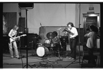 A band consisting of an electric guitarist, drummer, and euphonium player performs, as a person records them with a camera. Sound and lighting equipment are positioned around them.