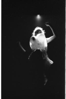 Low-angle shot of a woman with her arms raised, caught mid-motion. The lower half of her body is engulfed in darkness, while the upper part is illuminated by a stark spotlight. She wears balloons on her chest (perhaps imitating breasts) beneath a translucent top.
