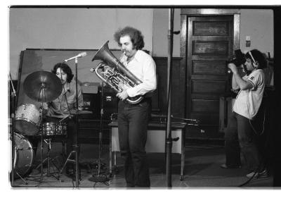 Two musicians play the drums and euphonium as a camera person records them. A microphone on a stand is positioned in front of the tuba player.