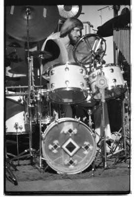 A bearded drummer with a bowl cut wears a pearl necklace, a light-colored v-neck top and white trousers. His white drum set fills up the majority of the frame, decorated with a small dream catcher and indigenous-inspired designs on the bass-drum. 
