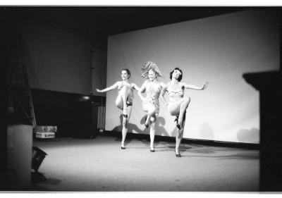 Three women wearing 50s-era bathing suits and kitten heels stand in a row with one leg up, cabaret-style. The women on each side have their arms outstretched, framing the one in the center, who wears a large zebra-print hat. They smile as they embody the pinup-girl performance. 
