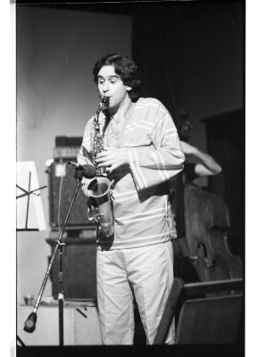 A dark-haired saxophonist glances to the side as he plays, seemingly nonchalant. He wears a striped, bell-sleeved top and  white trousers. A cellist and a pair of speakers are positioned behind him.
