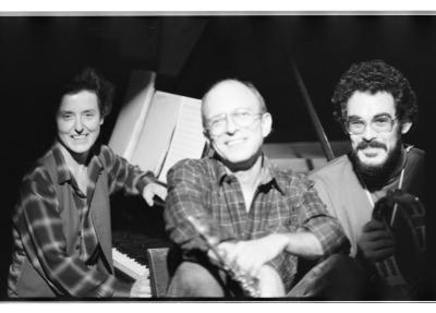 Three musicians smile broadly as they sit beneath a bright spotlight in front of a grand piano. In the center sits a man with a receding hairline, aviator glasses, and a plaid shirt. To his right is a younger man with dark, curly hair, glasses, and a beard. He wears a turtleneck and holds a tambourine. To the left a woman with short dark hair sits on the piano bench, wearing a plaid shirt and a vest.
