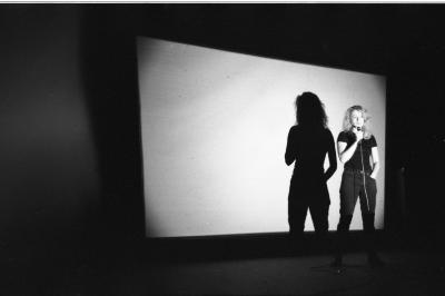  A woman stands while speaking into a wired microphone. She wears a dark t-shirt and jeans. Her shadow is cast on a projector screen behind her.