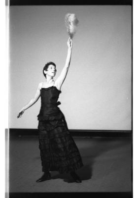 Marsha Paludan stands with their left hand raised, holding a large feather in the air, in front of a blank screen. Their hair is closely cropped to their face. They wear a long, formal gown with thin straps and layered detailing.