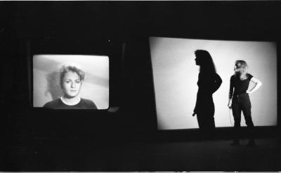 A woman stands with her hand on her hip and looks over her shoulder towards a monitor displaying a portrait of her. Her shadow is cast on a projector screen behind her. 