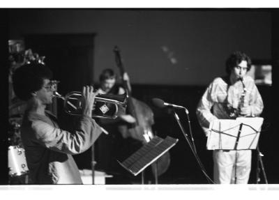 A trombonist with an afro and aviator-style glasses stands in side profile, reading off a score holder as he plays. Behind him, we can see the blurred figure of a short-haired cellist. To the far right stands a dark-haired saxophonist in a striped top reading off another score holder.
