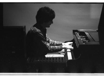 A high angle medium-long shot of a man playing the grand piano. There is a blank backdrop behind him, and in the foreground there is a computer interface instrument.