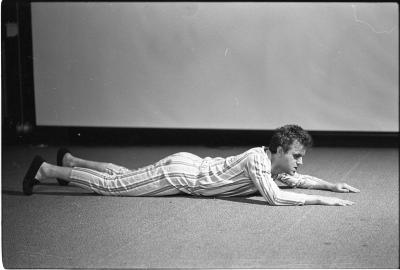 A man lying front-side down, parallel to the camera. His arms are bent and extended forward and his head is propped up. His hair is barbered, he is wearing a matching striped shirt and pants with slippers. In the background there is a projector screen. The image is in black and white.

