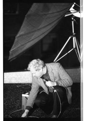 A light-haired man wearing a gray blazer, a dark shirt, and black trousers squats down, one hand holding a wired microphone and another touching the ground. He appears to be examining something on the floor. Behind him stands a large satellite dish.
