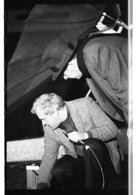 A light-haired man wearing a gray blazer squats down, one hand holding a wired microphone and another touching the ground, seemingly examining the floor. Another man wearing a dark coat and a fedora stands bent-over, also peering curiously at the ground. Behind them stands a large satellite dish.
