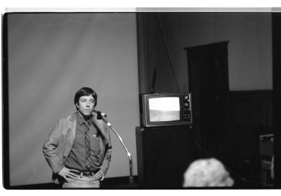 A dark-haired man wearing a blazer, dress shirt, and plaid tie stands with both hands on his hips, speaking into a microphone in the Grand Luxe Hall. To his left is an 80s-era TV displaying what looks like a horizon. We can see the back of one audience member’s light-haired head. 
