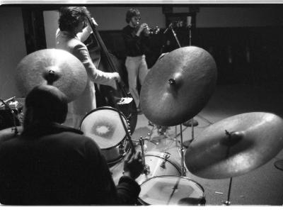 A high-angle shot behind the drummer, featuring three shining cymbals and a few drums. Other bandmates are featured behind the drum set; a cello and trumpet player. The image is in black and white.