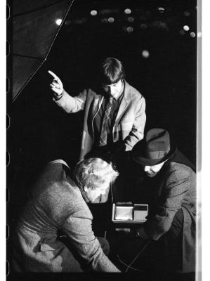 Three men in blazers gather around a satellite radio transmitter. A man in a fedora squats down to the right of the transmitter, facing a light-haired man reaching downwards. A dark-haired man wearing a plaid tie stands behind them, looking down at the transmitter as he points up at the edge of a satellite dish.
