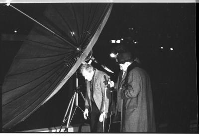 Three men wearing coats gather behind a satellite dish, seemingly inspecting its mechanisms. The man in the center holds a wired microphone in his hand. 
