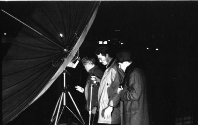 Side-profile of three men wearing coats gathered behind a satellite dish. The man closest to the dish is blonde, holding a hand up as if  to examine the tripod. The man in the center is dark-haired, with glasses and a mustache, holding a wired microphone. The third man wears a fedora and holds a bottle in one hand.
