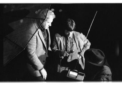 A translucent satellite dish vaguely obscures the side profile of a light-haired man wearing a blazer. To his left, a dark-haired man wearing a plaid tie holds a microphone aimed at a satellite transmitter with a huge antenna. In front of the transmitter sits a person wearing a fedora, their back facing the camera. 
