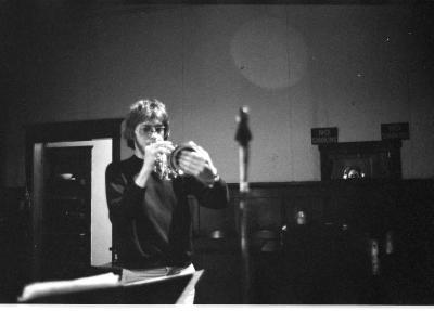 A medium-long shot of a man playing the trumpet. His left hand covers the front of the trumpet. He sports a 70’s mullet and aviator sunglasses. A door and wall decorations are featured in the background. The image is in black and white.
