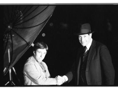 Two men pose beside a rooftop satellite dish, shaking hands and smiling at the camera. The man on the left is dark-haired, shorter, and wearing a light-colored blazer. The man on the right is taller, wearing a dark three-piece suit and a fedora. The sky is dark behind them.
