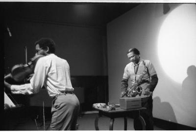 A man plays the violin in the foreground as he bends close to a music stand with some sheet music in front of him. Behind, another musician, carrying a saxophone plays a small percussion instrument that is placed on a table.