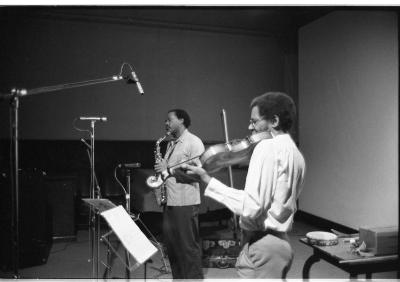 Two musicians perform together, one plays the saxophone and the other plays the violin. In front of them are three microphones and one music stand, and behind is a table with a small instrument on top of it. 