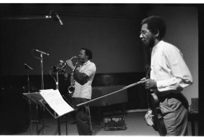 A musician with his violin in hand points his bow at a music stand with sheet music. Behind him another musician plays the saxophone, and has another hanging around his neck. In front of the two artists are three microphones resting on stands at various heights.