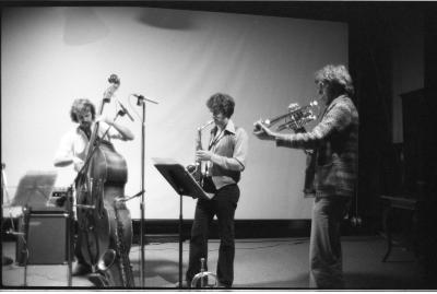 A trio of musicians play instruments, from left to right they play the cello, saxophone, and trumpet. Several pieces of music equipment surround them. The background features a large blank backdrop. The image is in black and white.