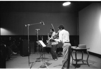 A long-shot of two musicians perform together, one plays the saxophone and the other plays the violin. In front of them are three microphones and one music stand, and behind is a table with a small instrument on top of it. The audience's presence is insinuated through crossed legs on the left side of the image.