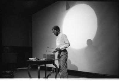 A musician plays a small percussion instrument that rests on a table. He holds two xylophone mallets, in either hand. A bright spotlight is directed towards him, leaving a glowing circular impression on the backdrop behind him.
